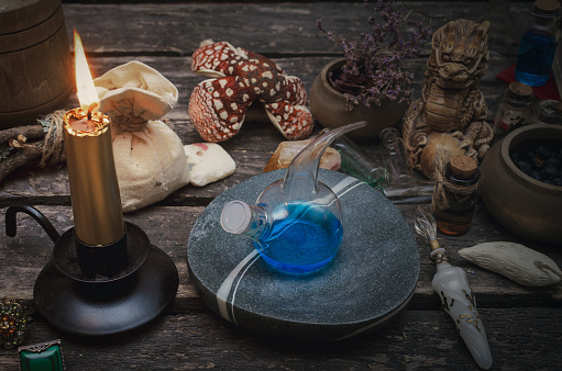 Ein mystischer Tisch mit einer brennenden Goldkerze, einem blauen Trank in einer Glasretorte, einer kleinen Drachenstatue, getrockneten Kräutern und einem Stoffbeutel auf Holzbrettern.