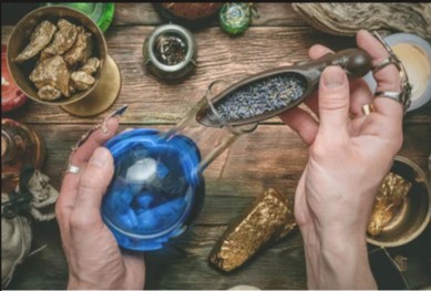 Eine Person mit Ringen an den Fingern gießt getrockneten Lavendel aus einer Holzschaufel in eine leuchtend blaue Glaskugel auf einem rustikalen Holztisch mit Gold.
